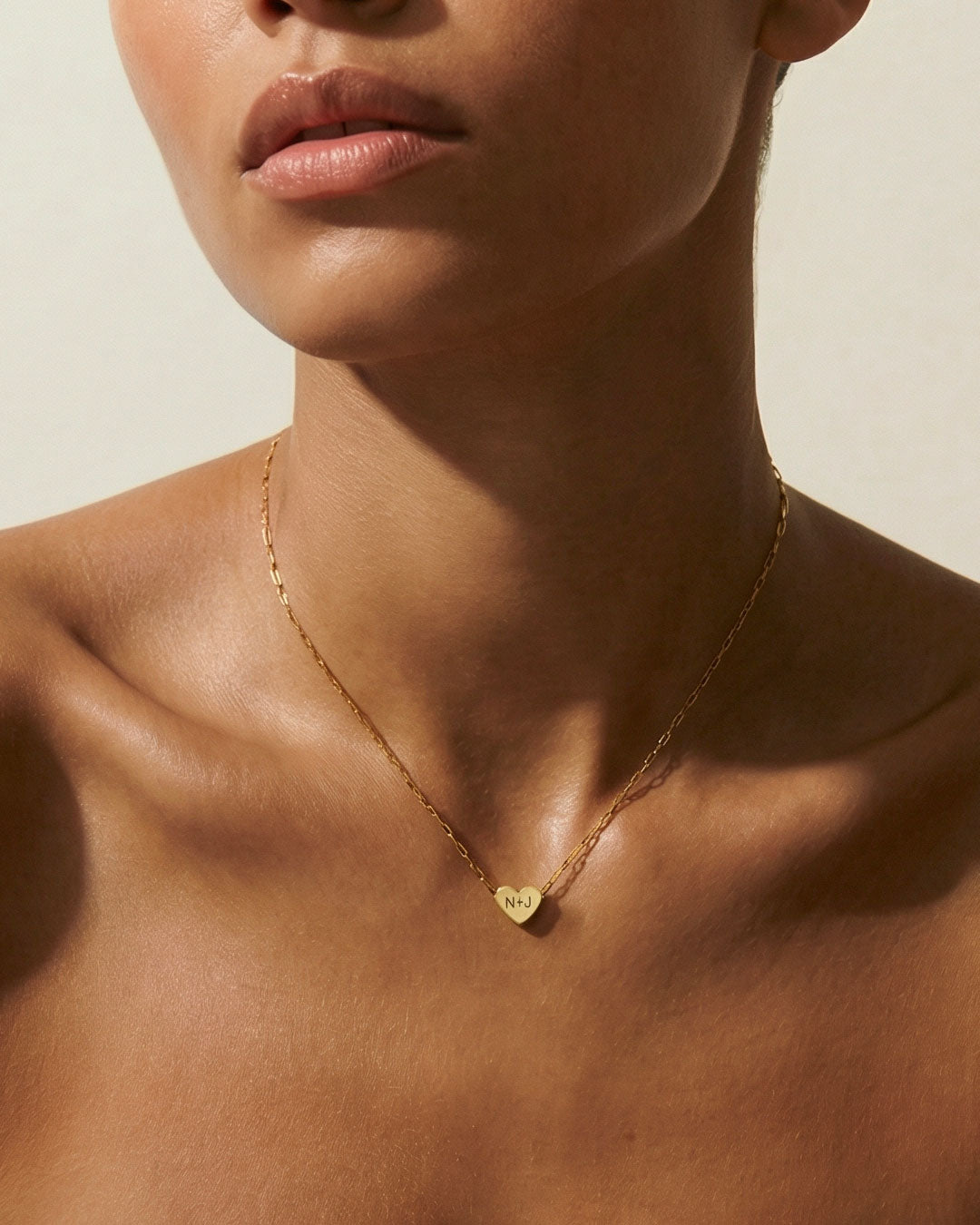 Close-up of a person wearing a gold necklace with a heart-shaped pendant on a neutral background