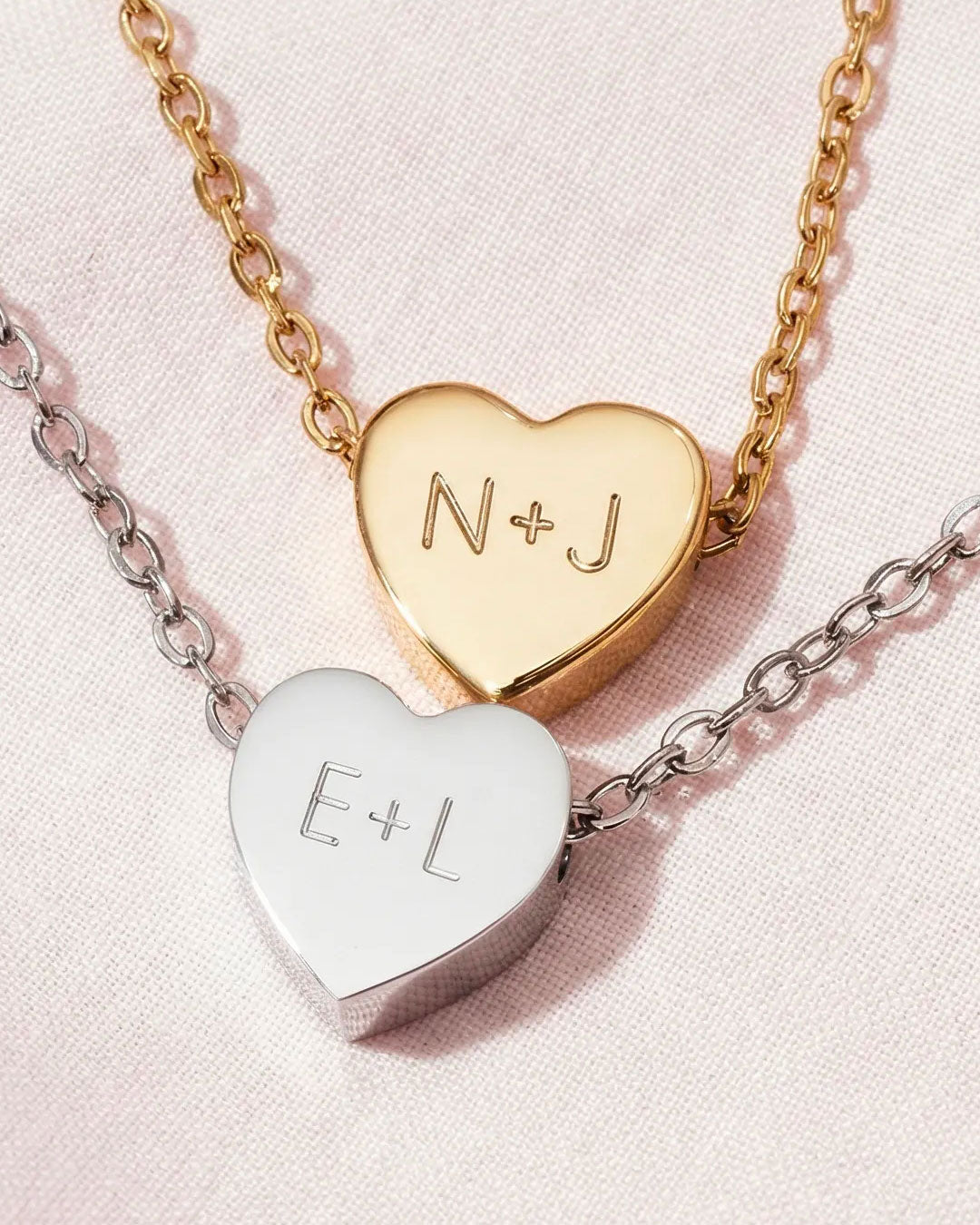 Two heart-shaped pendants on chains with engraved initials on a pink background